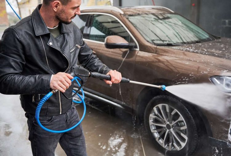 Young man washing car on carwash station outdoor. Handsome worker cleaning automobile, using high pressure water.